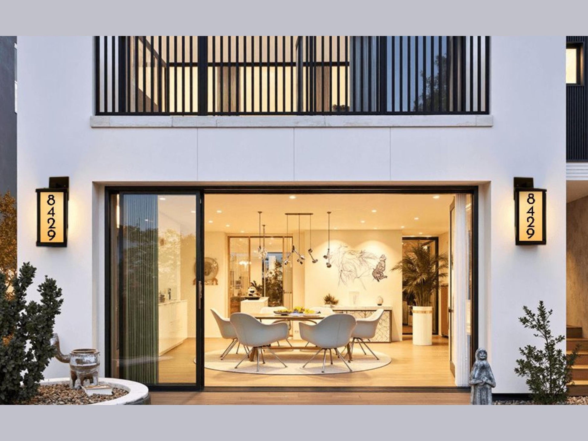 a modern, open-concept living space with a dining area, featuring a glass wall that provides a view of the interior. The space is illuminated by pendant lights hanging from the ceiling, creating a warm and inviting atmosphere. The room is decorated with various plants and artwork, adding to the overall aesthetic.