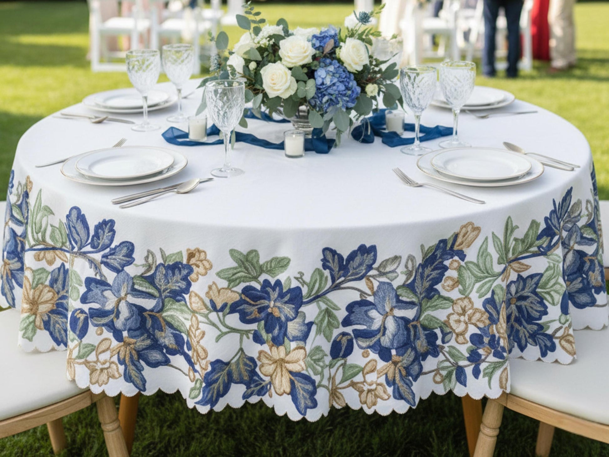 a beautifully set outdoor dining table with a white tablecloth featuring a floral pattern in blue, green, and gold colors. The table is set with plates, silverware, and wine glasses, creating an elegant and inviting atmosphere.