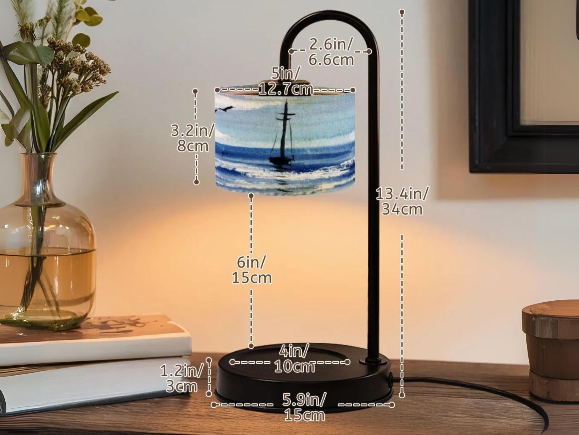 a black lamp with a sailboat design on the lampshade, placed on a wooden table. The lamp has a black metal base and a black metal stand. The lampshade measures 2'6" in height and 6" in width. The lamp is accompanied by a vase with flowers and a book, creating a cozy and inviting atmosphere.