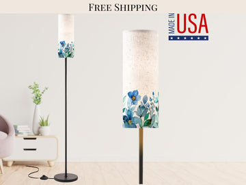 a modern living room with a tall floor lamp that has a white lampshade with a floral design. The lamp is placed on a wooden floor, and there is a white table with a plant and a vase on it. The background of the image is a white wall with a logo that says "FREE SHIPPING MADE IN USA".