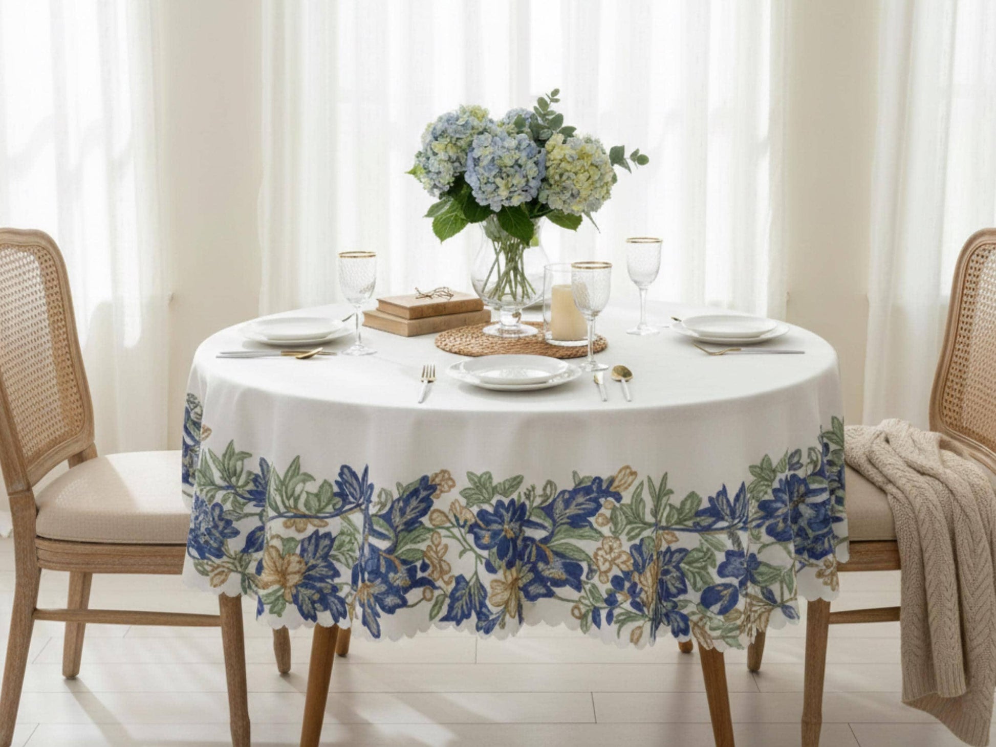 a cozy dining scene with a round table set for two, featuring a floral tablecloth, plates, silverware, and a vase of blue hydrangeas. The table is positioned in front of a window, allowing natural light to illuminate the setting.